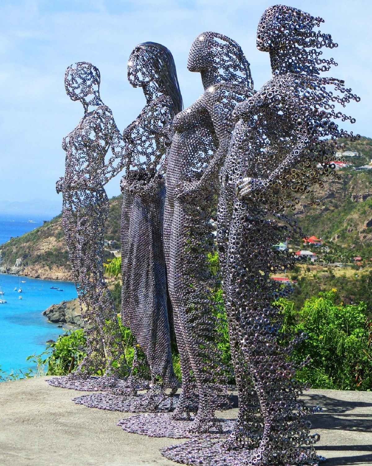 Four human sculptures made from stainless steel nuts by Jean Martin, standing on a concrete platform overlooking the sea in Saint Barthélemy. The figures gradually fragment toward the back, as if wind is blowing through them.
