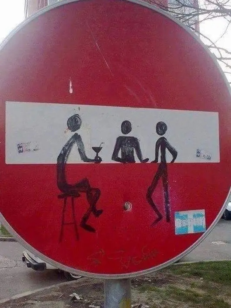 Modified “Do Not Enter” sign showing stick figures sitting and drinking at a bar drawn across the white band of the sign, turning the traffic symbol into a humorous nightlife scene.