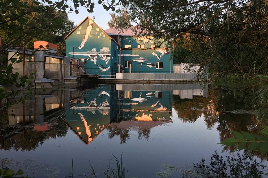 The turquoise mural by artist Ray Bartkus in Marijampolė, Lithuania, beautifully reflected in the calm canal water at sunset. The upside-down design on the building wall transforms into a harmonious upright scene of swimmers, rowers, and swans in the water’s reflection. The warm golden light and surrounding trees enhance the serene atmosphere of the setting.