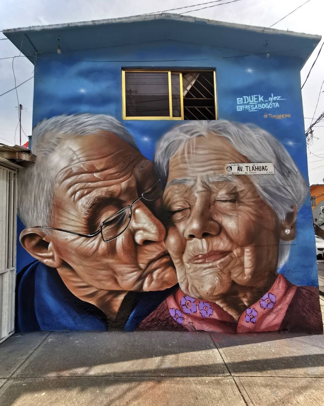 Realistic mural of an elderly man kissing an elderly woman on the cheek, painted by Duek and Fresa Bogota on a blue house in Tláhuac, Mexico.
