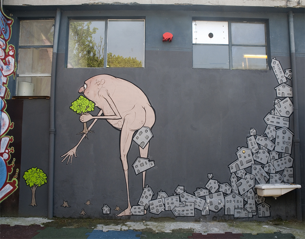 A thought-provoking mural titled "Cagacemento" by NemO’s, located in Milan, Italy. The artwork features a naked, hunched humanoid figure with elongated limbs, clutching a small green tree. Behind the figure, a trail of gray, block-like houses spills out from its body, symbolizing urban sprawl and environmental destruction. In contrast, tiny vibrant trees grow at the edges of the mural, emphasizing the conflict between nature and relentless human expansion. The stark, minimalist design on a gray wall underscores the ecological message of preserving greenery amidst urbanization.