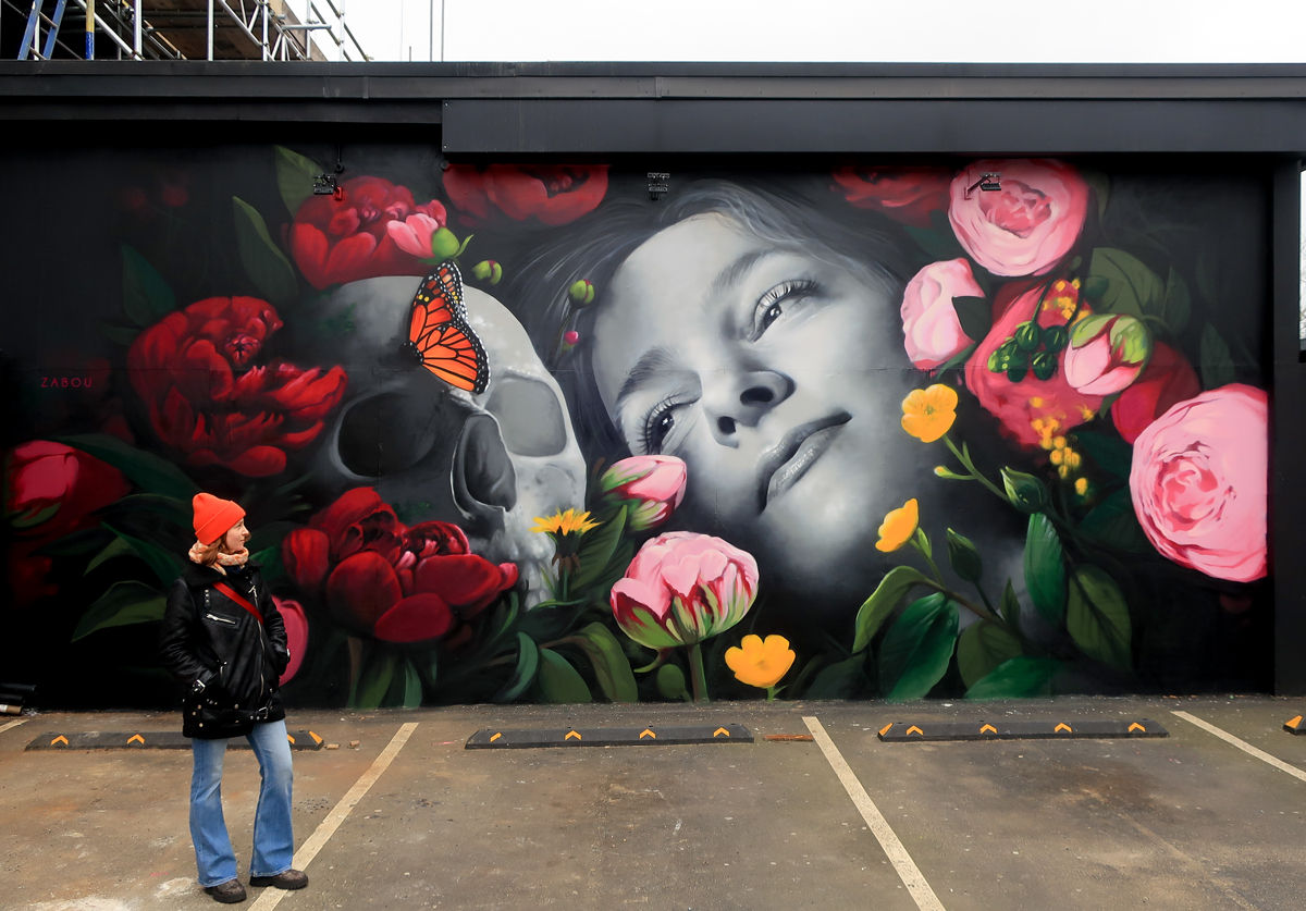 ZABOU mural in London showing a grayscale woman’s face beside a skull surrounded by red and pink flowers and an orange butterfly, photographed with a woman in an orange beanie standing in front.