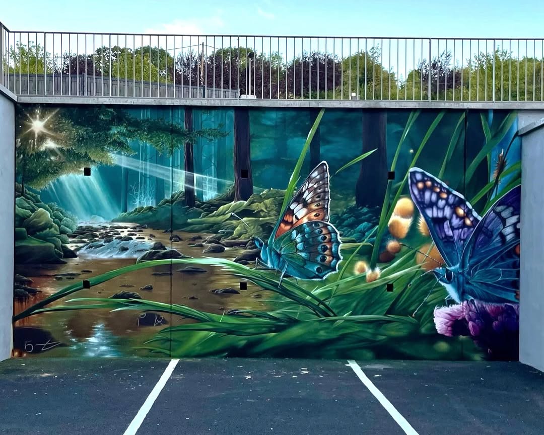 Mural by Dege in Le Puy-en-Velay, France, depicting two oversized butterflies in tall grass beside a forest stream lit by sunbeams.