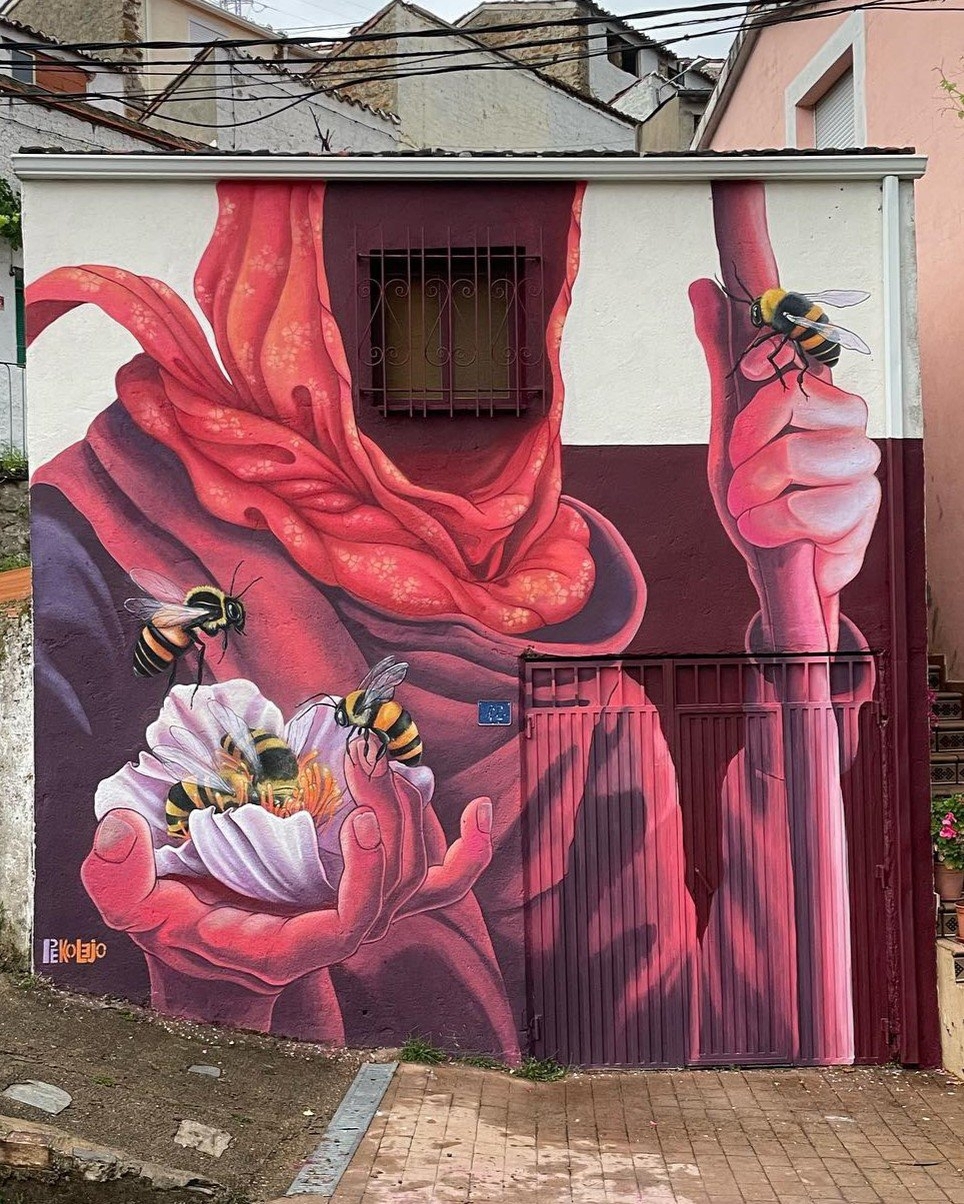Vibrant mural on a house wall of a red-robed goblin holding blooming white flowers with bees collecting pollen, painted in warm pinks and reds.