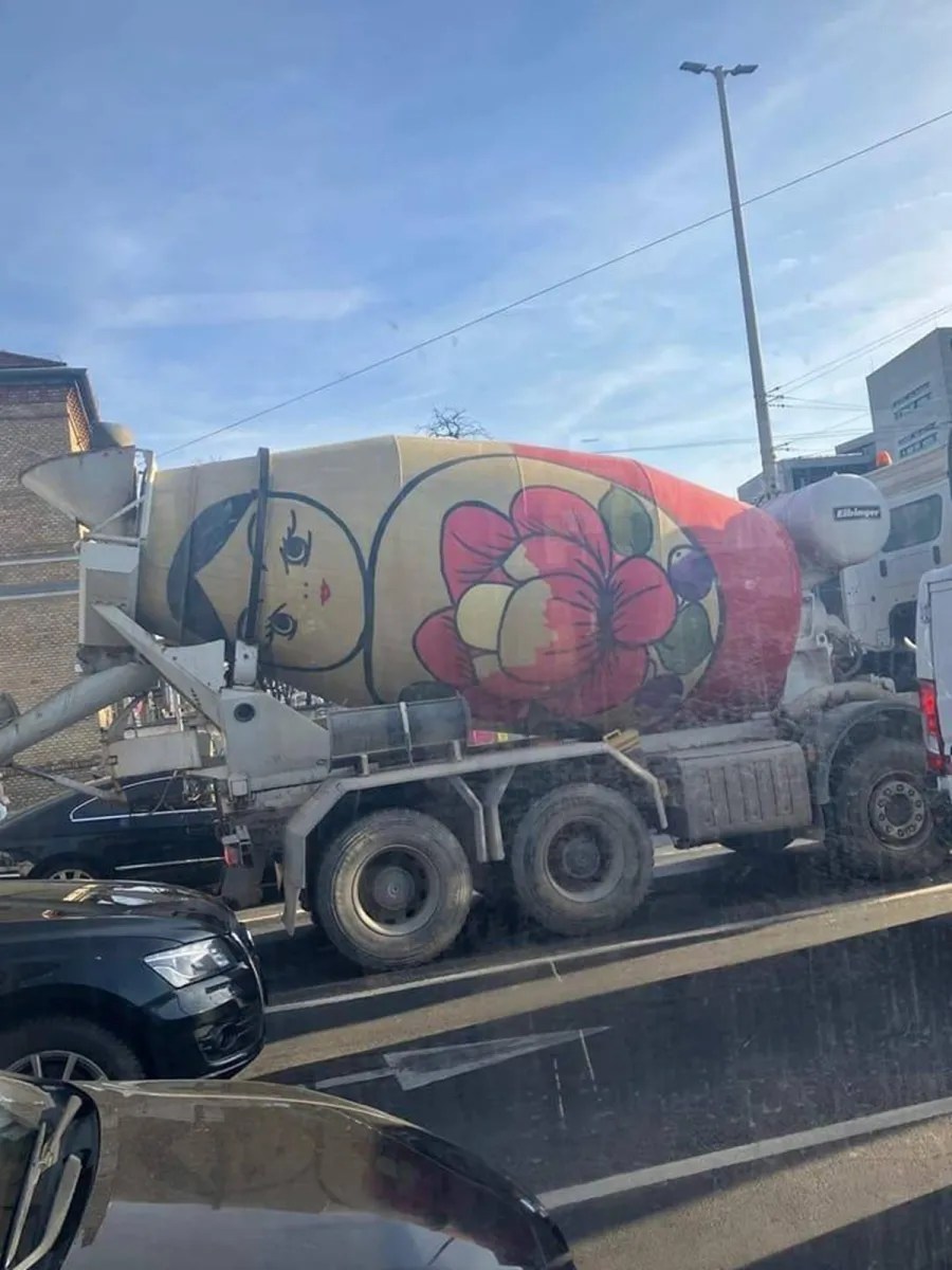 A cement truck in Budapest painted to resemble a Matryoshka doll, also known as a Russian nesting doll. The truck's rotating drum features a stylized face with black hair, red lips, and a large floral design with vibrant red and yellow hues, mimicking the traditional folk art patterns of Matryoshka dolls. The truck is seen stuck in traffic, blending industrial function with playful artistic expression.