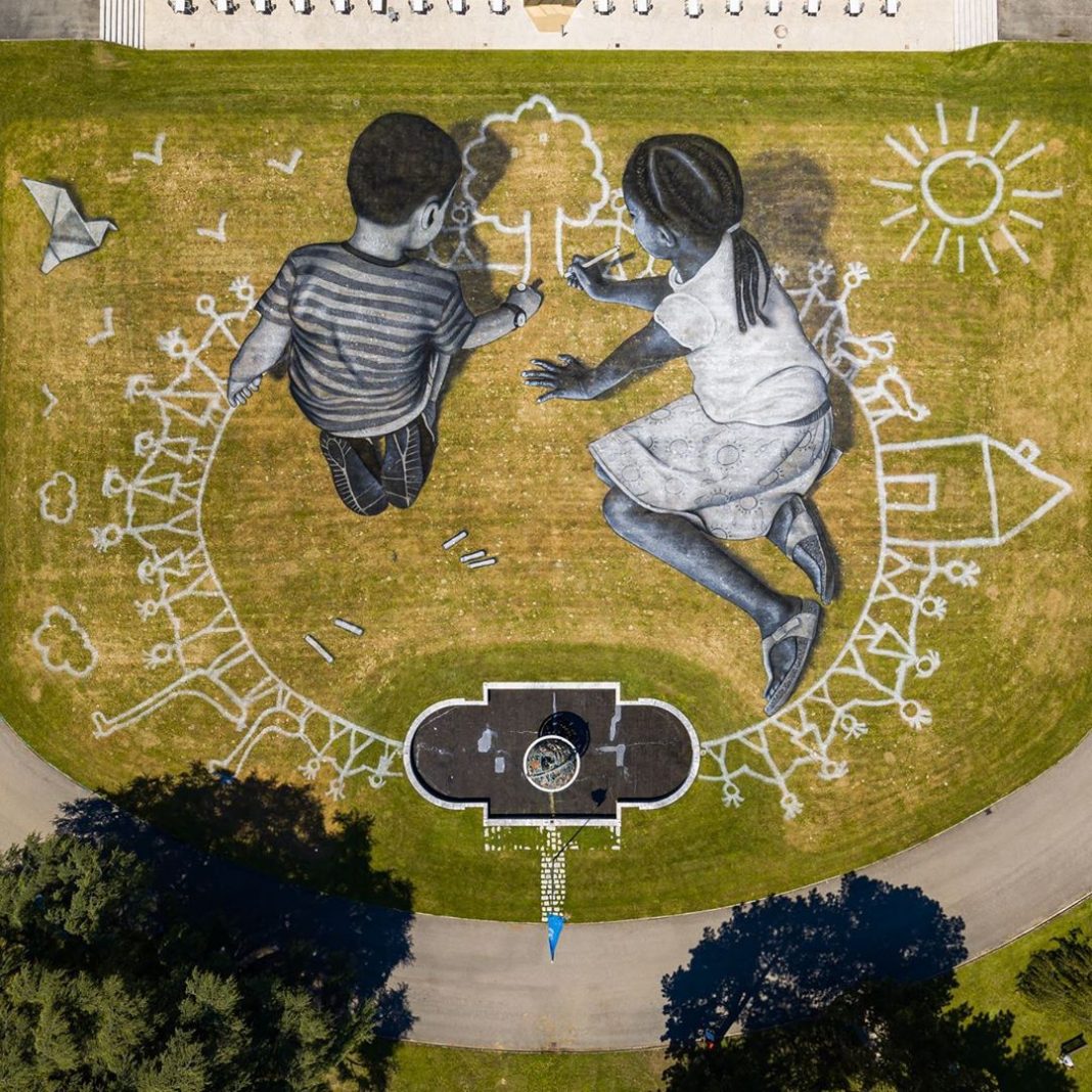 Monumental land art by Saype in Geneva, Switzerland, featuring two children drawing on a massive stretch of grass titled World in Progress.