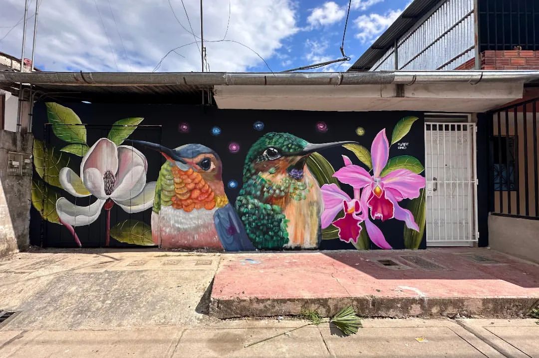 Vibrant hummingbird and flower mural by Safe in Moyobamba, Peru, glowing with color against a dark background.