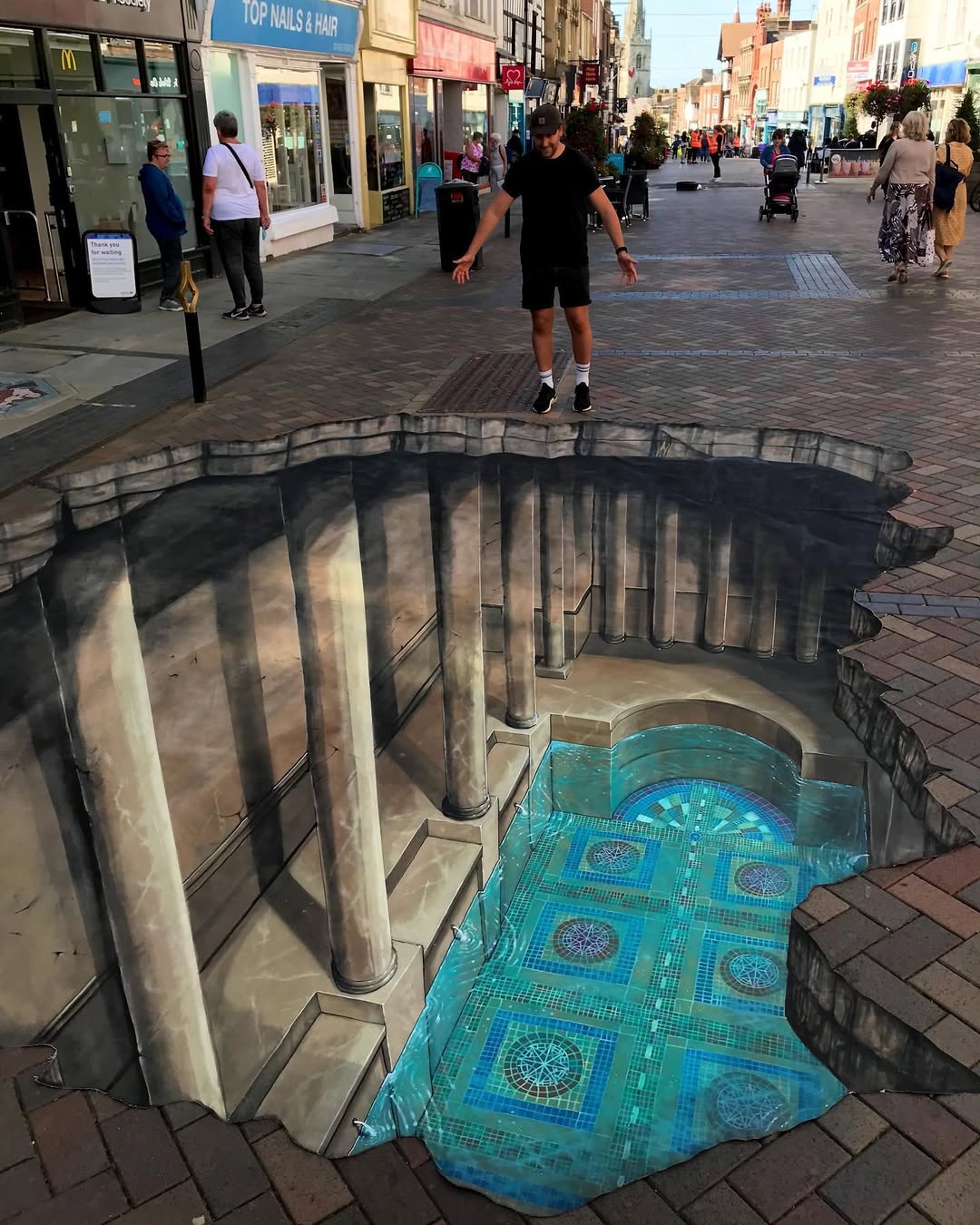 A 3D chalk street artwork showing a man standing above an illusionary pit in the middle of a pedestrian street. Inside the hole, classical columns and a pool with blue mosaic tiles are painted to appear real.