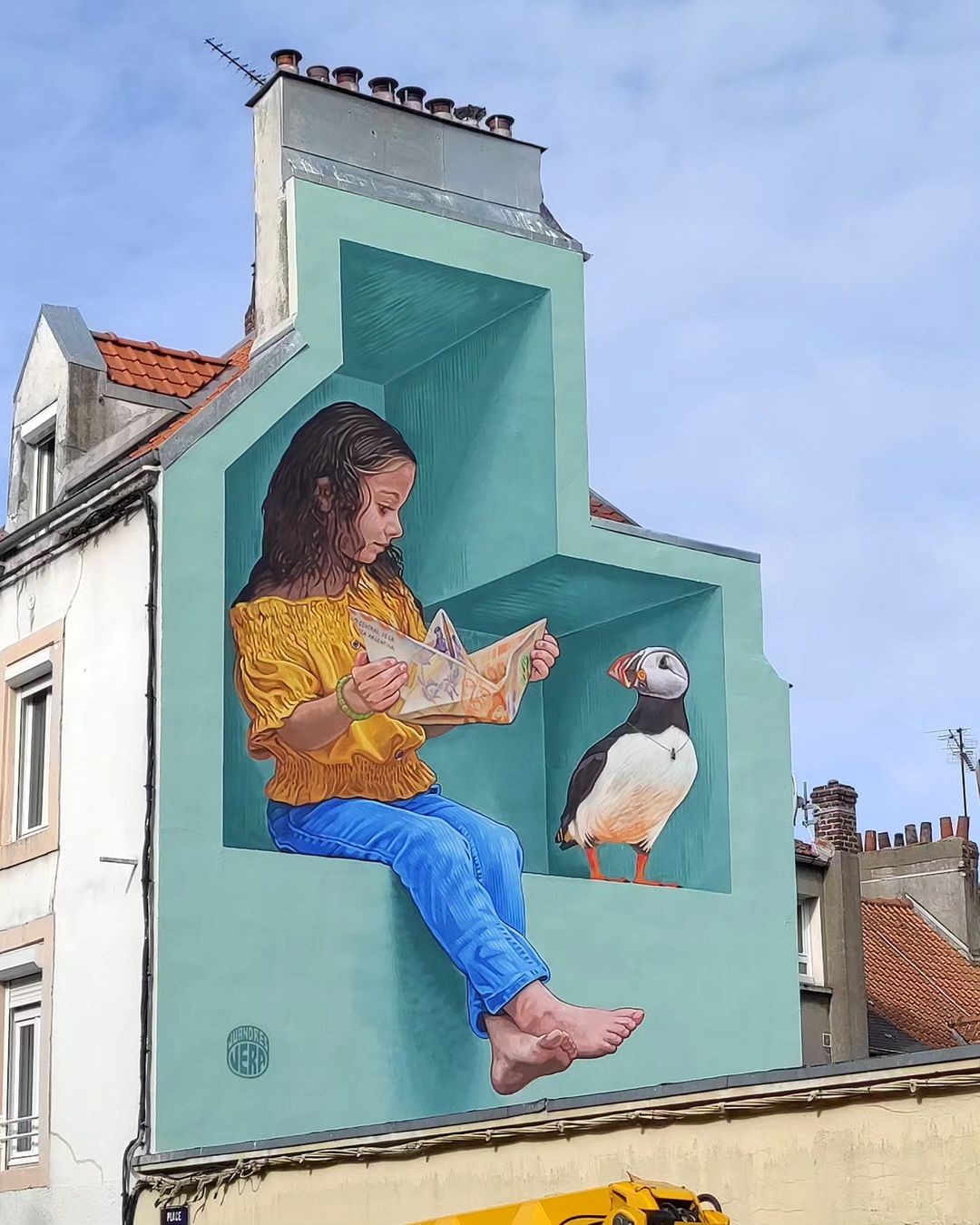 A mural of a young girl reading a book next to a puffin, painted inside a turquoise cube on the side of a building. The cube creates a 3D illusion using shading and perspective, making it appear as an architectural extension.