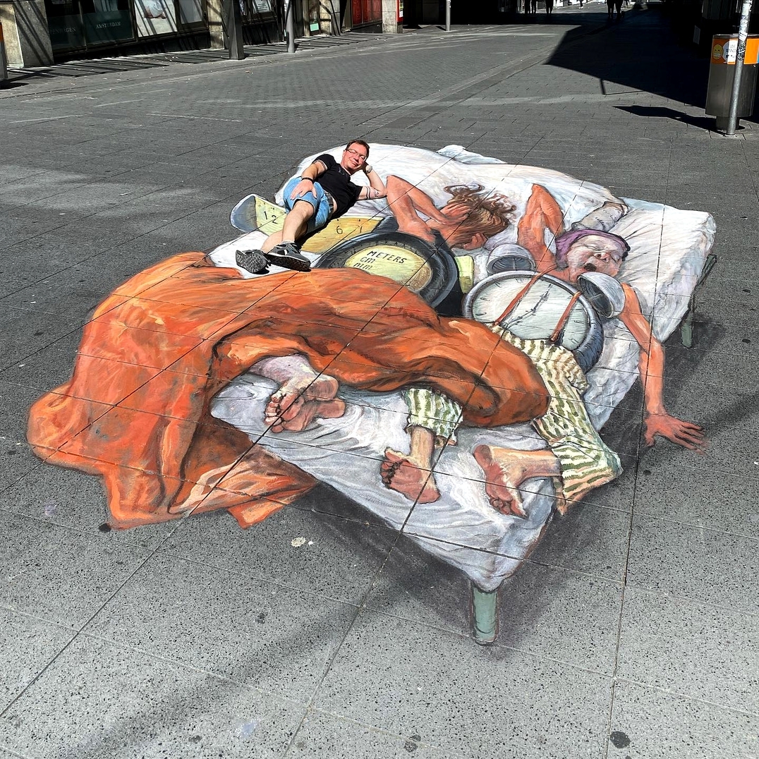 A 3D chalk artwork on a city street shows two elderly cartoonish men sleeping in bed. A real person lies among them, integrated into the illusion. The mattress and characters are painted with shadows to appear realistic.