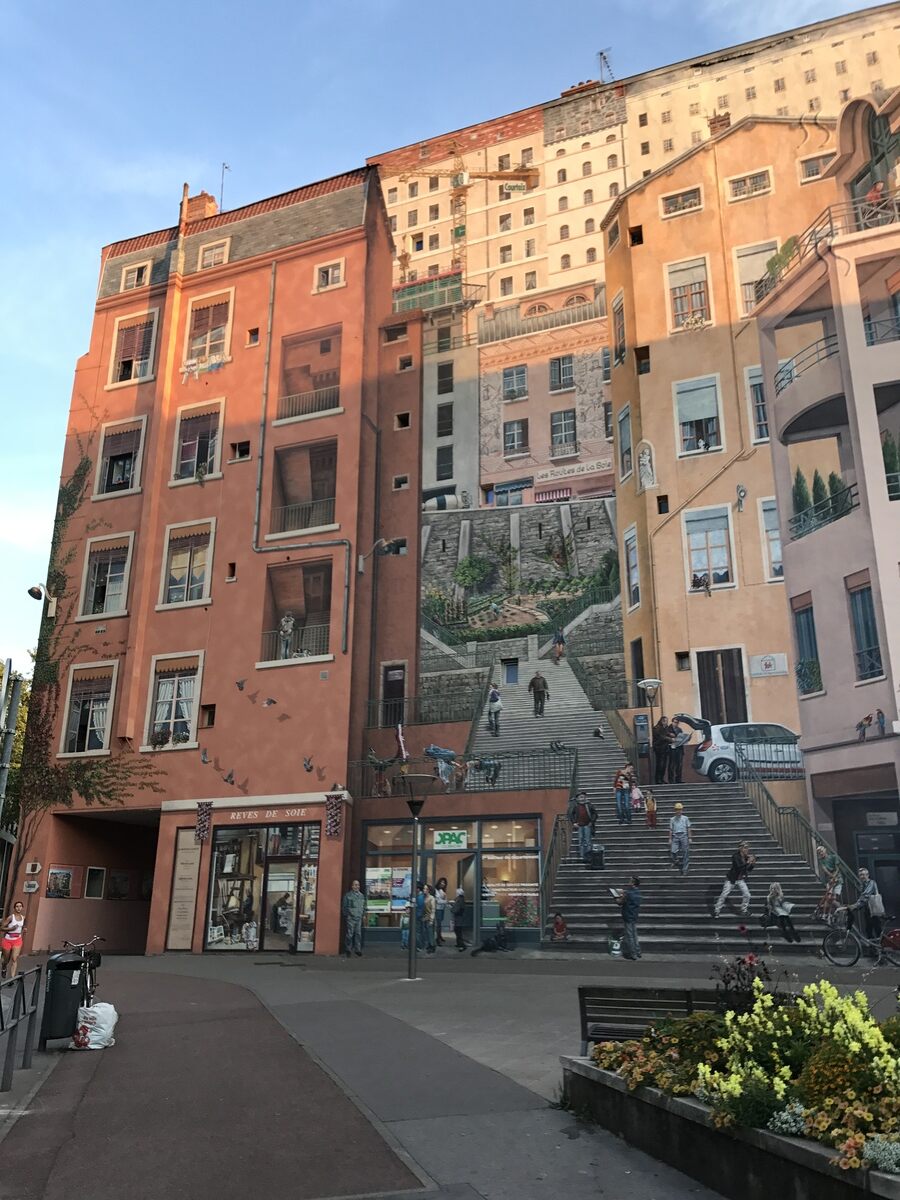 A wide-angle view of the 'Wall of the Silk Weavers' mural in Lyon, France, capturing the detailed trompe-l'œil artistry that transforms the building facade into a lively urban scene. The mural spans multiple stories, depicting buildings with balconies, windows, and a central staircase filled with painted figures going about their day. Shops and storefronts at the ground level appear three-dimensional, blending seamlessly with the real street environment. Flower beds in the foreground add color, complementing the warm hues of the mural under soft evening light. The mural pays homage to Lyon’s rich history in the silk weaving industry, bringing vibrant life and charm to this urban space.