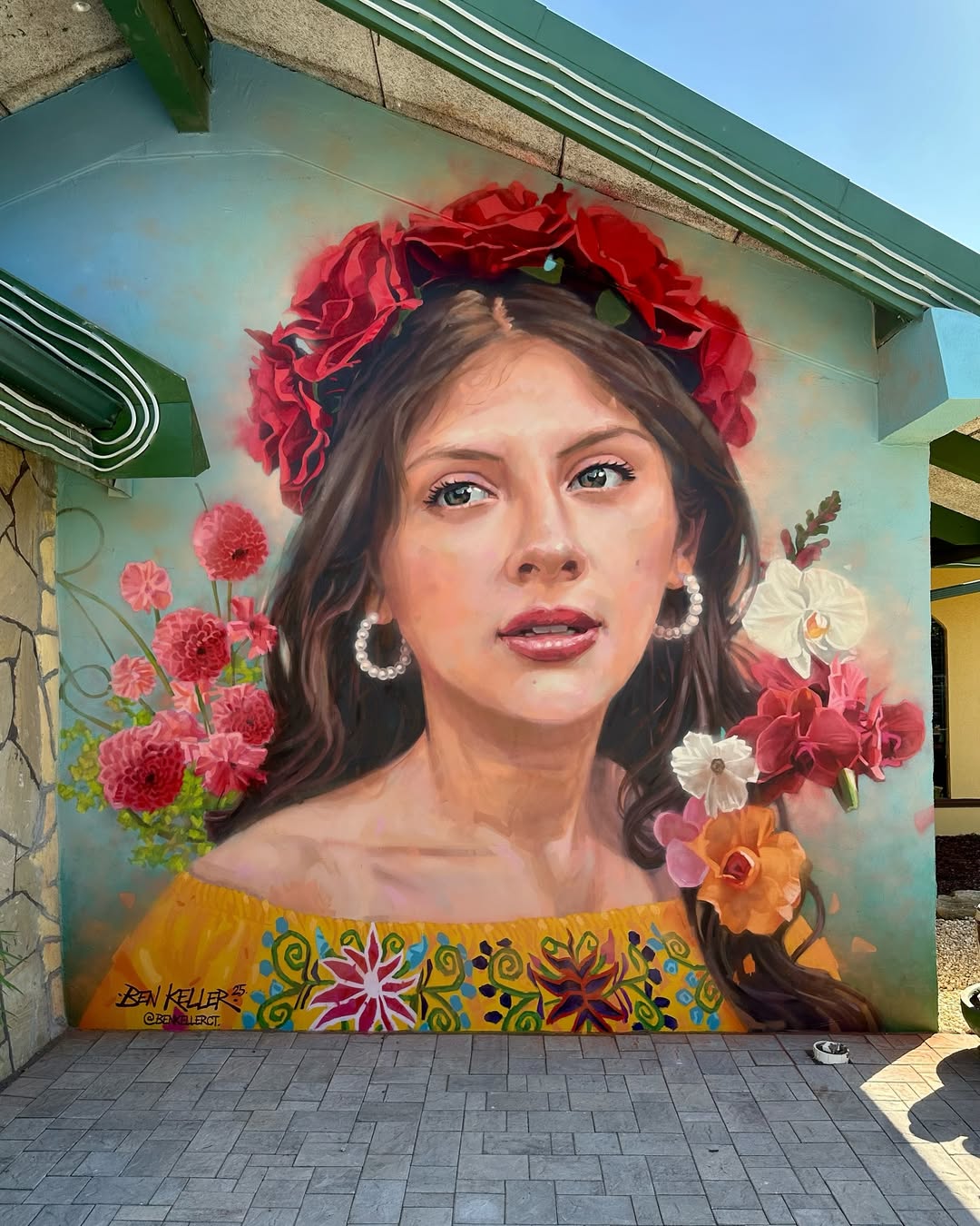 Mural of a woman with long dark hair adorned with red roses and pearl earrings, painted by Ben Keller in Charlotte, North Carolina. She wears a bright yellow dress with floral embroidery, surrounded by vivid flowers on a teal background.