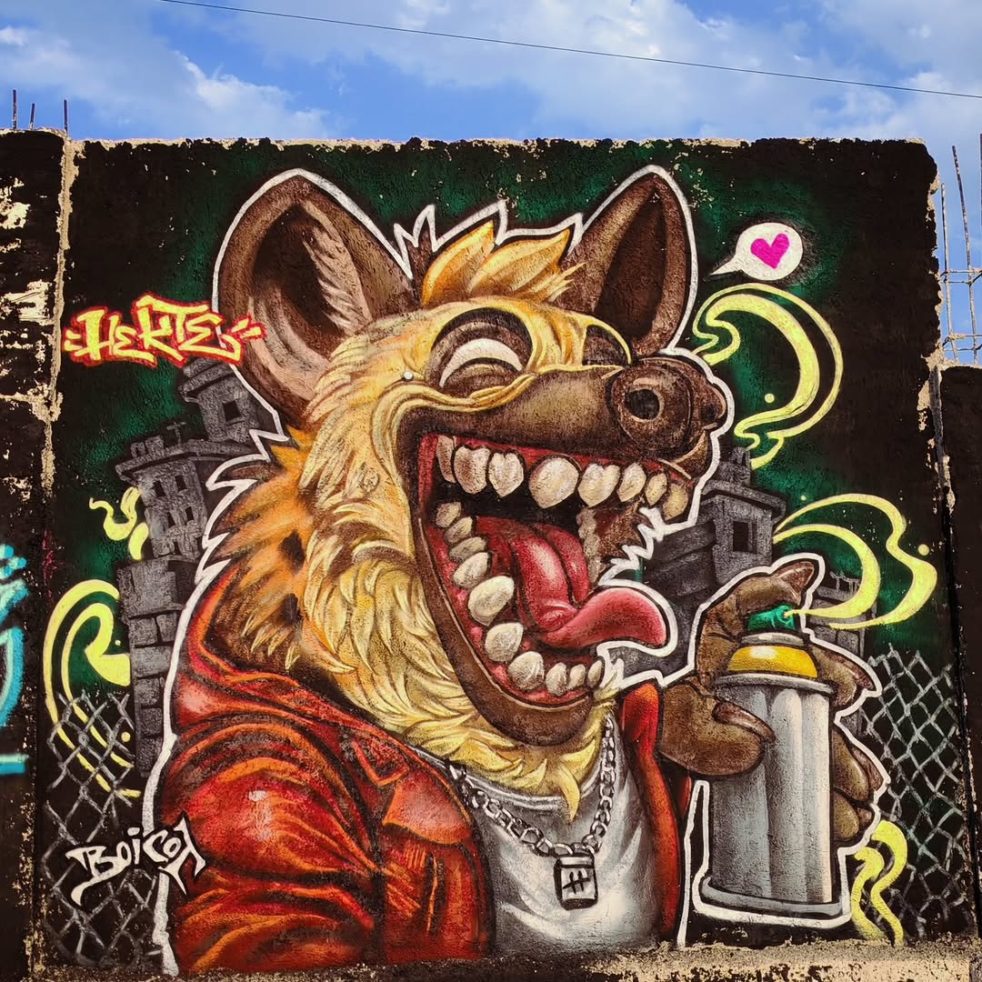 Colorful graffiti-style mural of a laughing hyena holding a spray can, created by Jean Hekte in San Juan Teotihuacán, Mexico. The background shows crumbling buildings and neon green accents against a black wall.
