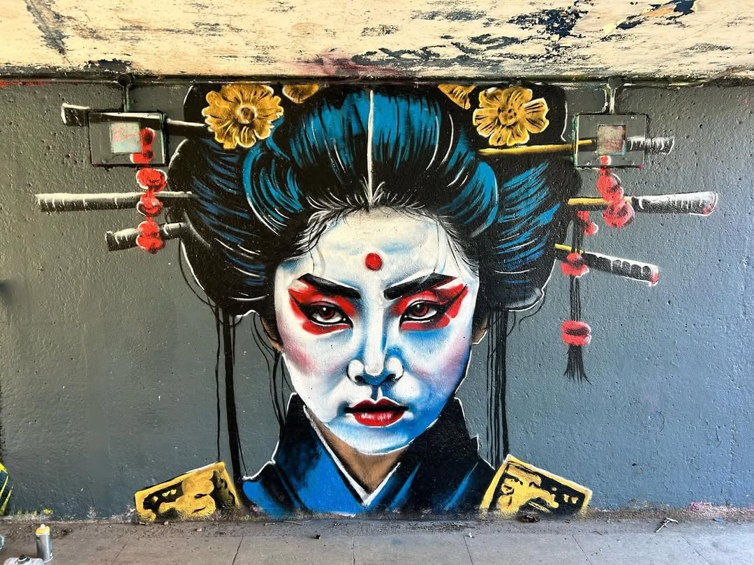 Street art portrait of a geisha with a red mark on her forehead, intense red and white makeup, and swords in her hair, painted by Steve Dixon in Hull, UK. Her expression is stern against a grey concrete wall.