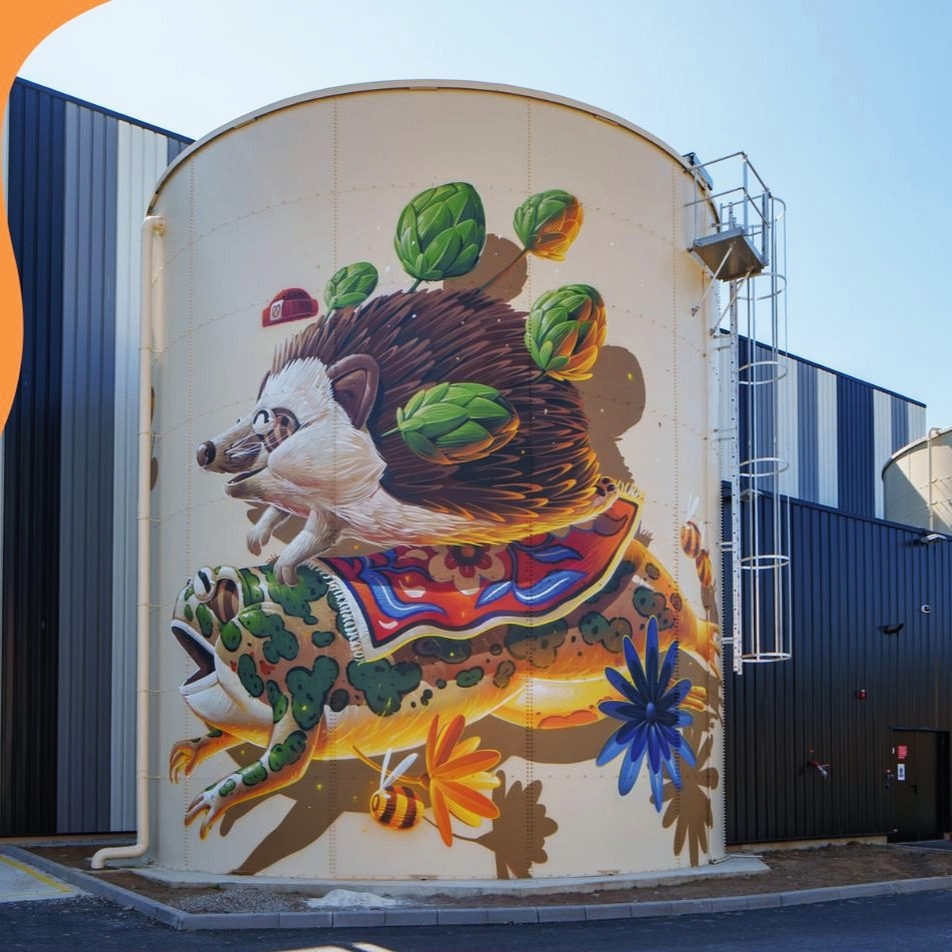 Mural of a hedgehog riding a frog with lettuce-like leaves on its back, painted on a white silo by STOM500 in Duppigheim, France. A bee and colorful flowers swirl around the duo in motion.