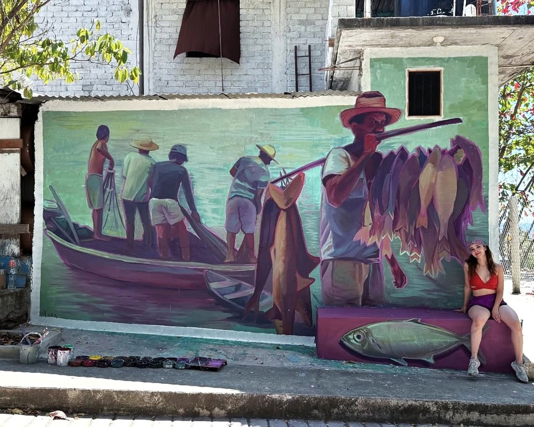Mural of fishermen unloading boats and a vendor with a pole of hanging fish, painted by YUDA in Puerto Escondido, Mexico. A woman sits beside the mural, aligned with a painted bench featuring a realistic fish.