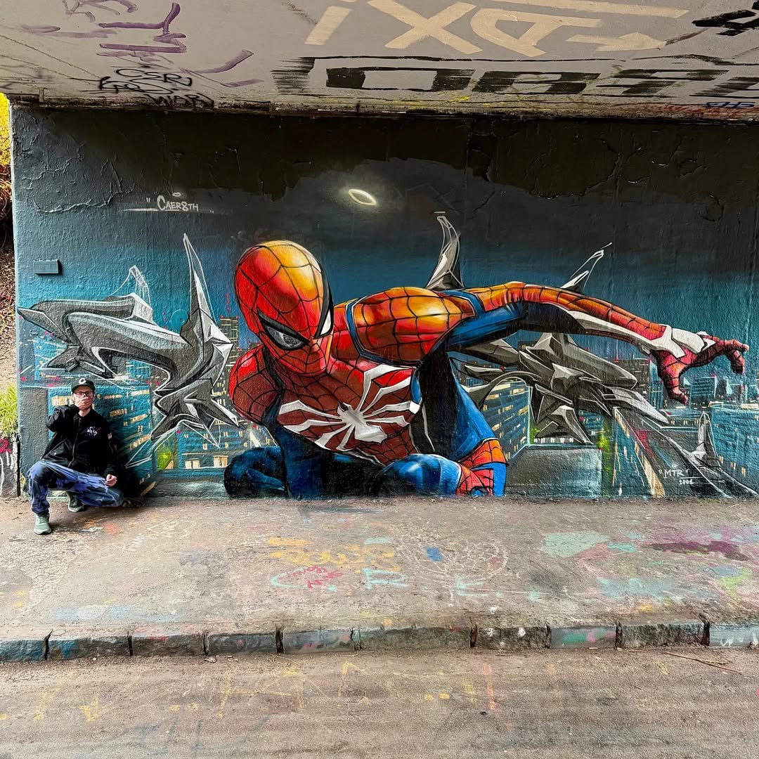 Mural of Spider-Man crouching with one hand outstretched, painted by Vladimír Hirscher in Prague, Czech Republic. The background features a night city skyline with a glowing halo above Spider-Man’s head.