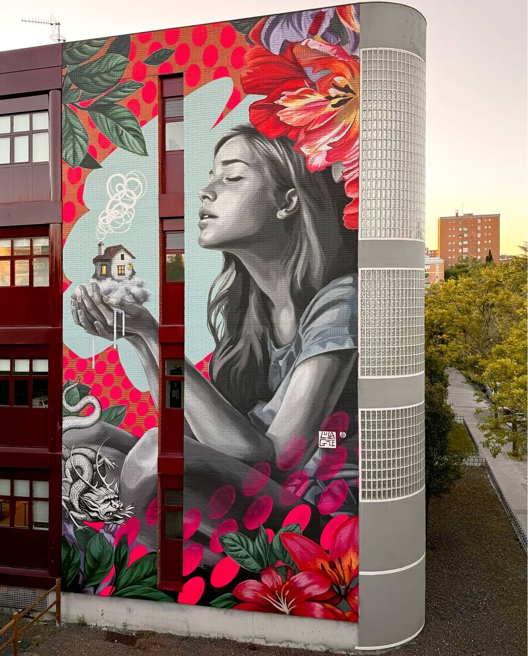 Large mural in Madrid by Lula Goce showing a grayscale woman with long hair holding a miniature house emitting white smoke, surrounded by red and pink flowers, a dragon, and colorful polka-dot and leaf patterns.