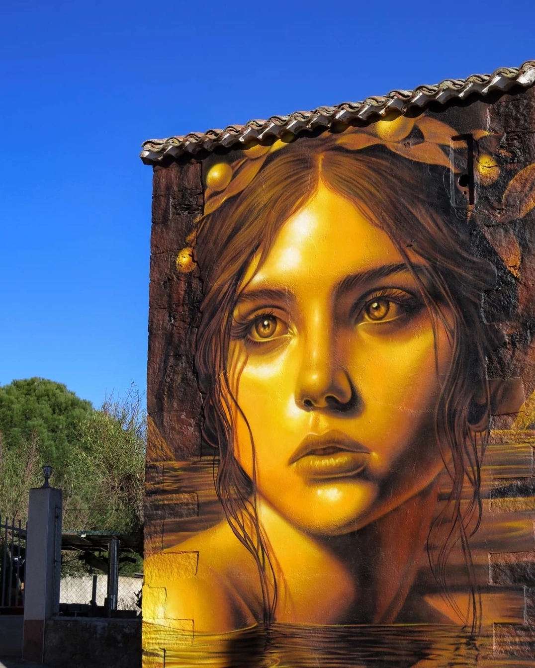 Golden-toned mural of a young woman’s face in Cinco Villas, Spain by Moxaico, with lemon fruits above her head and water reflections at the bottom, set against a bright blue sky.
