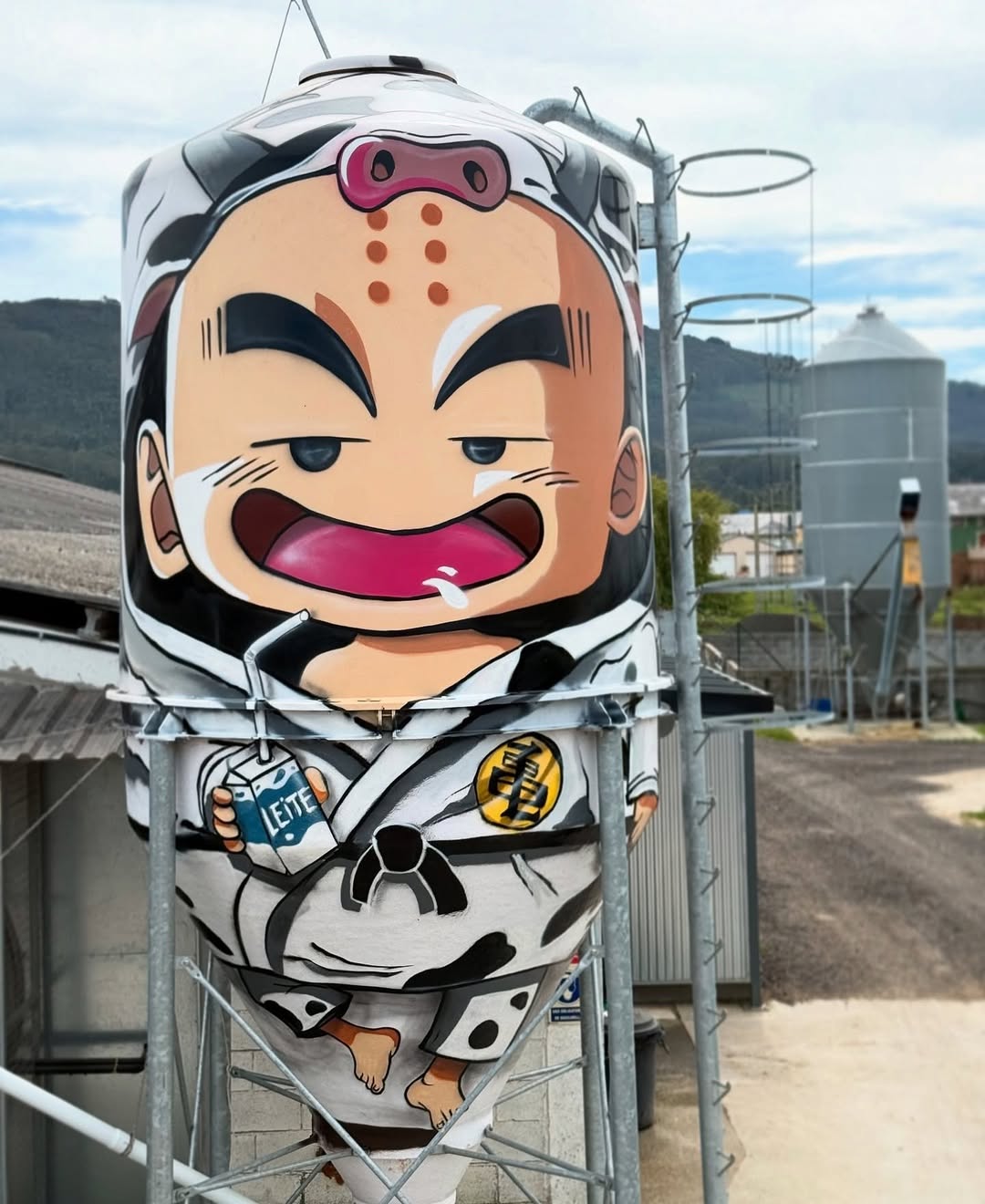 Cartoon mural on a milk silo in Ribadeo, Spain by TRECE TRAZOS, showing a martial arts character in a gi, holding a milk carton with a wide grin and raised eyebrows, painted on a cone-shaped tank.