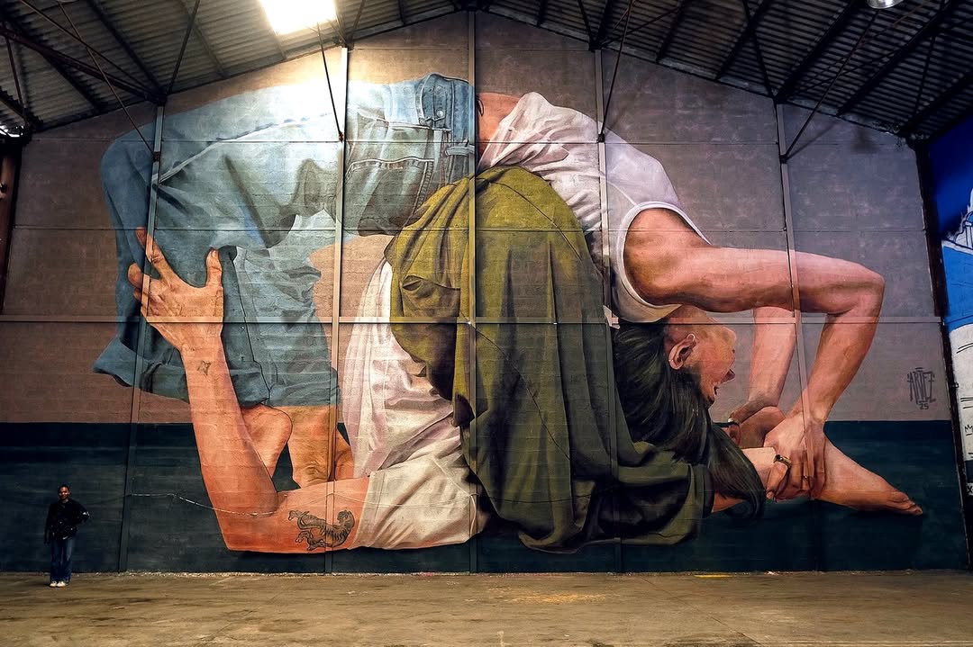 Mural titled “Simple Acrobatics” by Artez in Bourgoin-Jallieu, France, showing two large intertwined figures in a yoga pose painted on an indoor warehouse wall, with one upside down in a stretch, in photorealistic style.