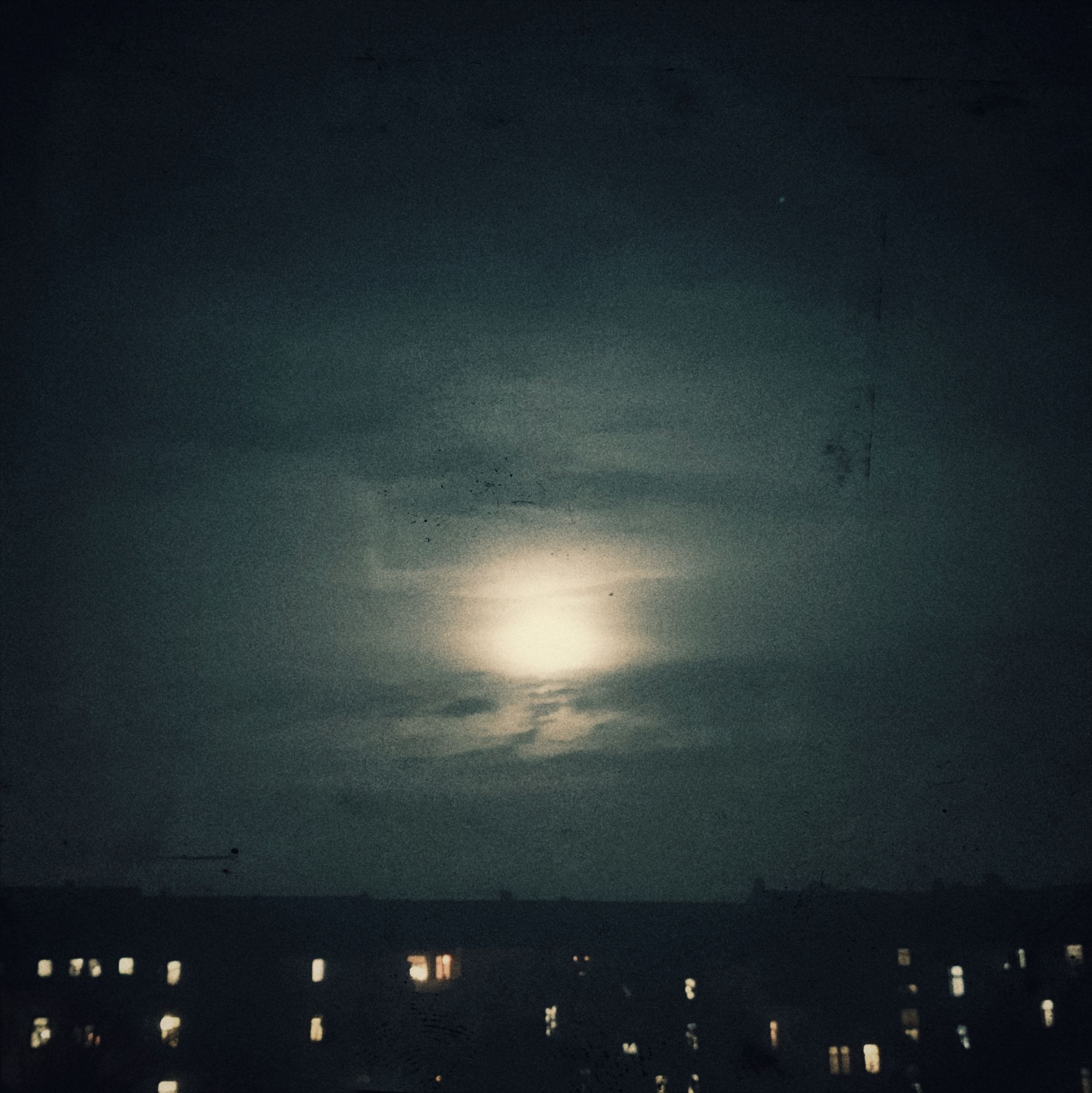 Häuserzeile, erleuchtete Fenster, Himmel. Mond in Wolkenschleier.