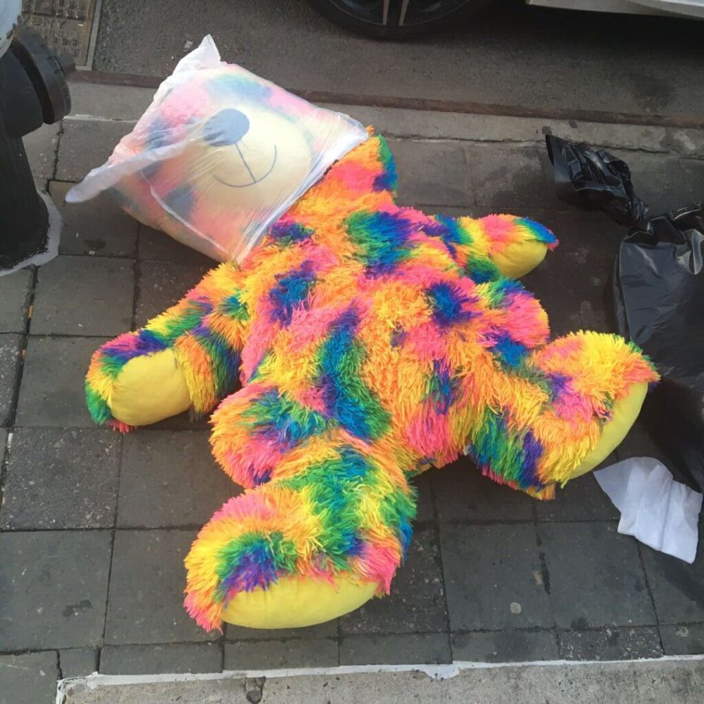 A massive teddy bear with rainbow fur lies down on the curb with a white translucent garbage bag tightly fitting on its head.