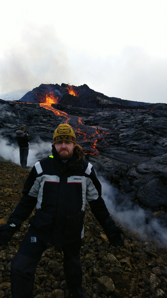 Rysiek, in heavy weather sailing outfit, a knit cap with "Ryś" on it, and the volcano and some nice lava river in the background.