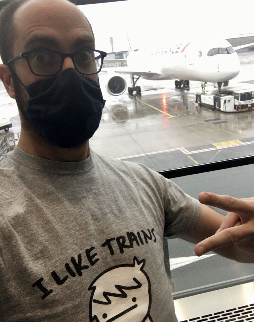 Selfie of myself wearing a tee-shirt with the ASDF Movie’s “I like Trains” song title and character inside an airport terminal, looking outside where a passenger jetplane is parked at a gate.