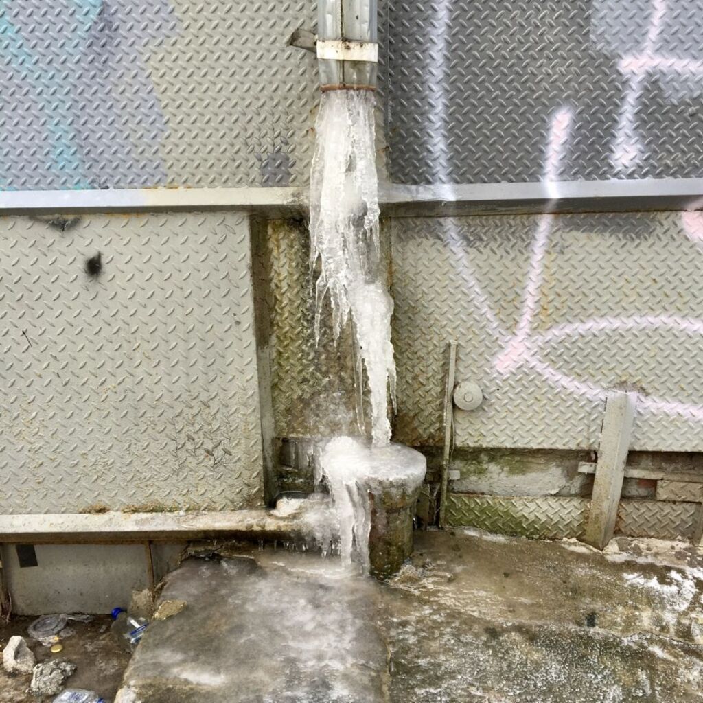 A drain pipe on a street wall is cut at waist height and a formation of ice pours from it, spreading on the sidewalk.