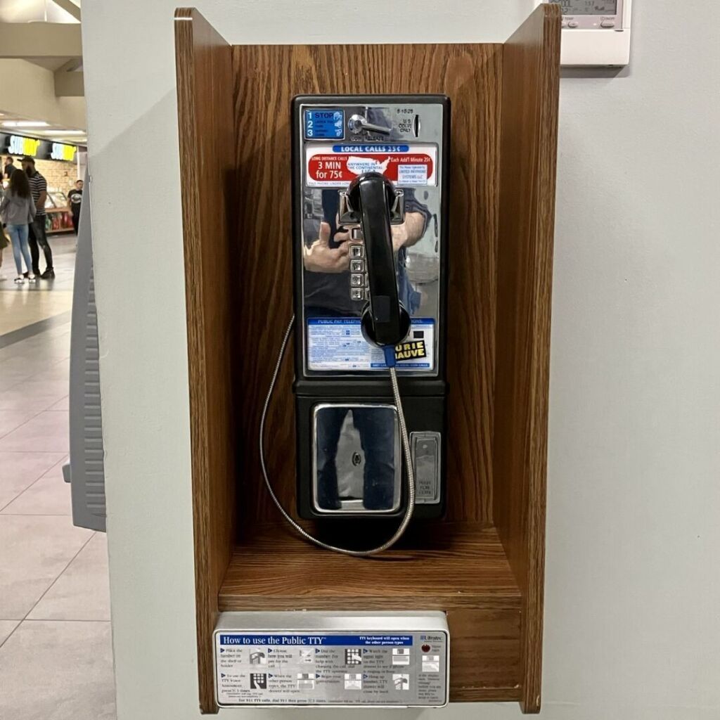 A chromed payphone in a wooden enclosure on a white wall.