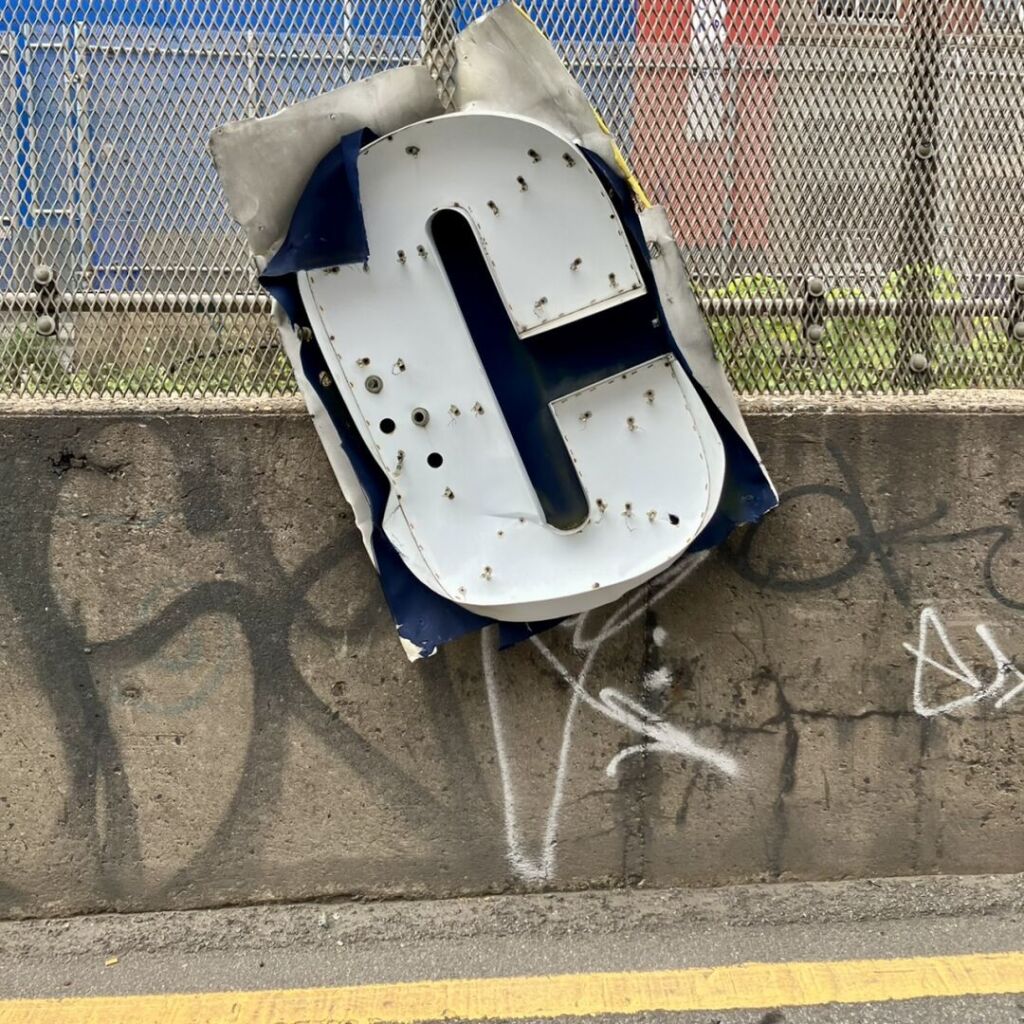 A large white bent metal marquee letter C is hanging off a roadside fence above a graffiti-laden concrete barrier.