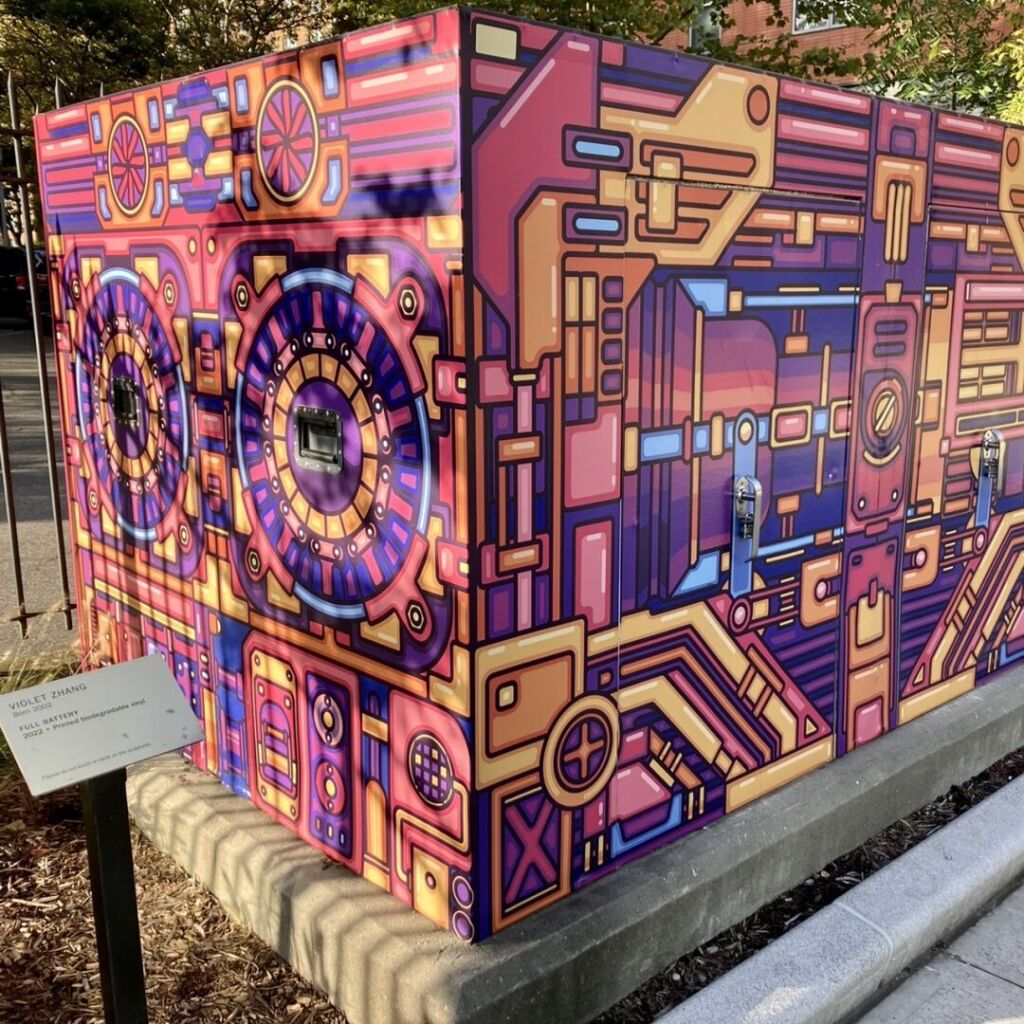 A large outdoors electrical cabinet on the sidewalk is covered in colorful vinyl with a pattern evoking the interior of a computer case.