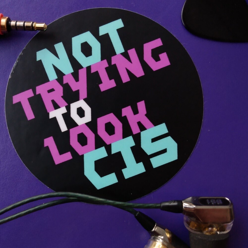 Three inch circle "Not Trying To Look Cis" vinyl sticker on a notebook with earbuds and guitar pick for scale.