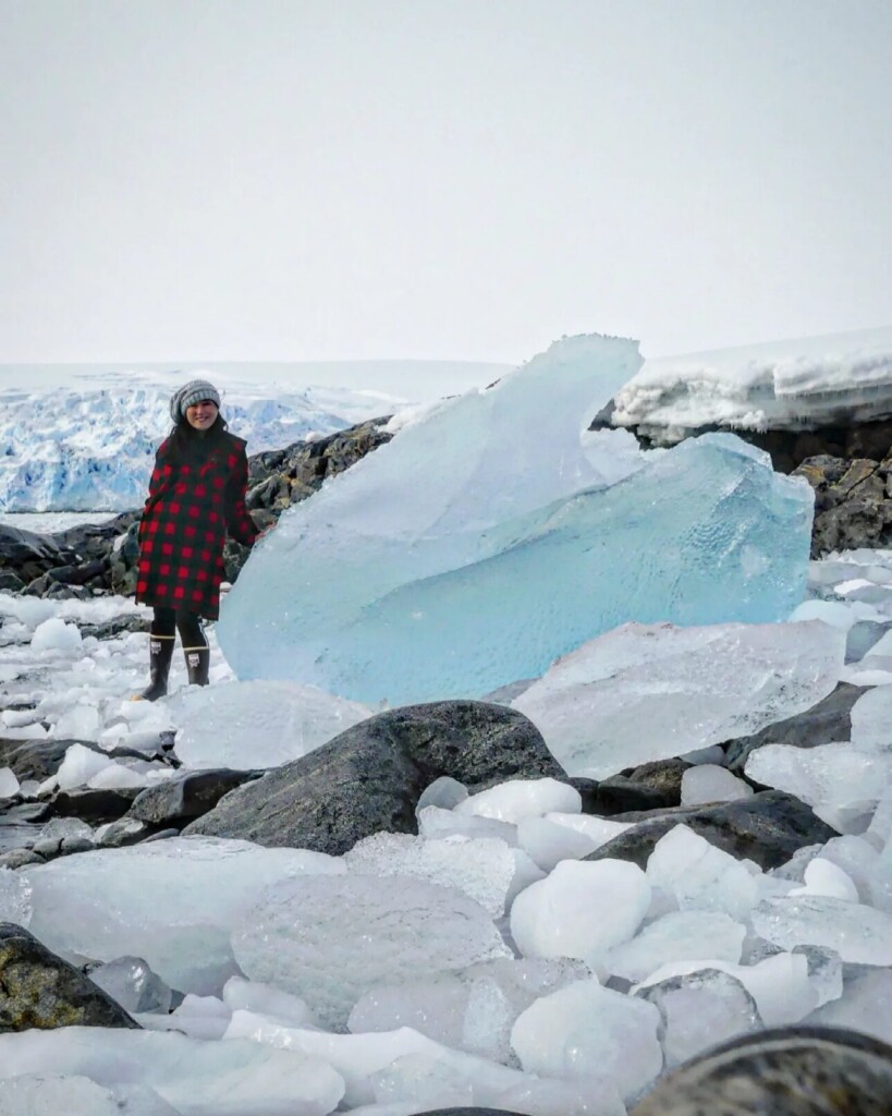 165 centimeter person stands on rocky shore next to large clear glacial blue ice chunk as tall as her, and three times as long.