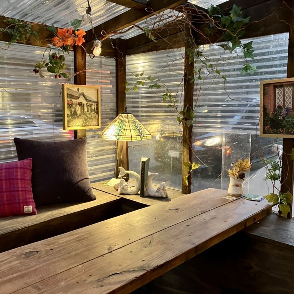 A restaurant street wooden shack is adorned with books, framed artworks, pillows and a table lamp with a stained glass lampshade projecting a warm light around.