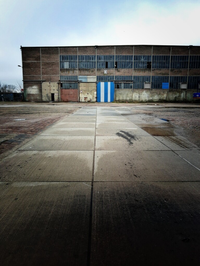 View of an abandoned and dilapidated factory. Some windows are broken. There's a large blue-white-striped door and a sort of road leading up to it.