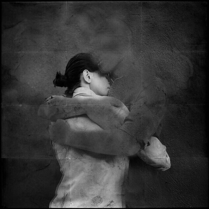 A moody grey image of a woman wih her back to the viewer being hugged or held by a pair of ghostly grey arms emerging from the misty background. 