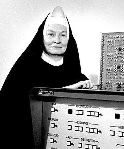 Black and white photo of a smiling nun in a habit standing next to a large bank of old computer equipment.