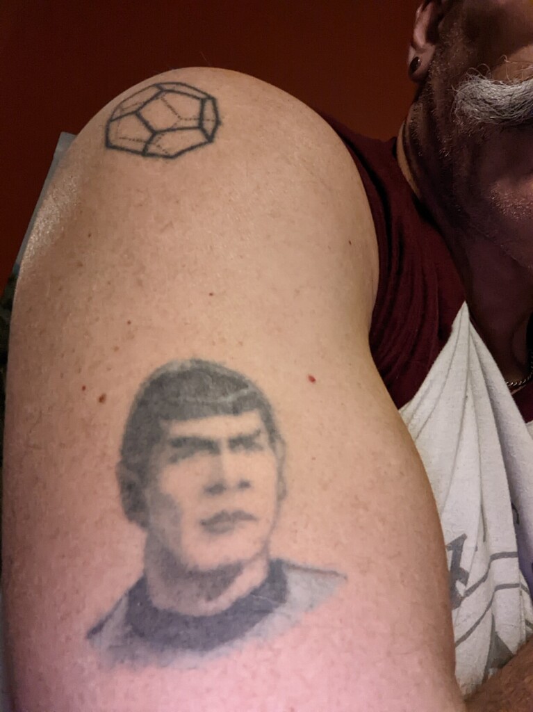 Arm with faded tattoo of a bust of Spock, and a dodecahedron, and a man with a long moustache in the background 