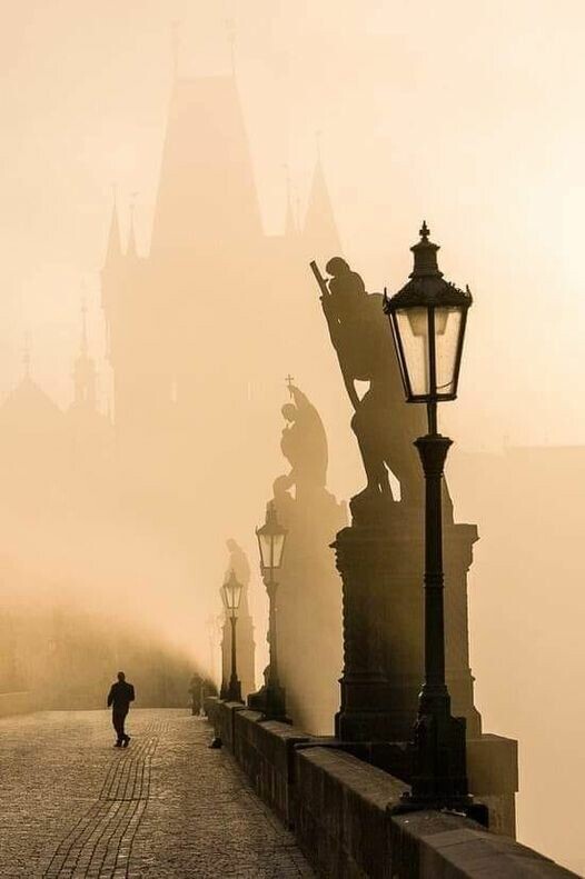 Prag, Karlsbrücke im Nebel. Ein Mann geht über der Brücke, man sieht die Skulpturen auf der Brücke und die Strassenlaternen.