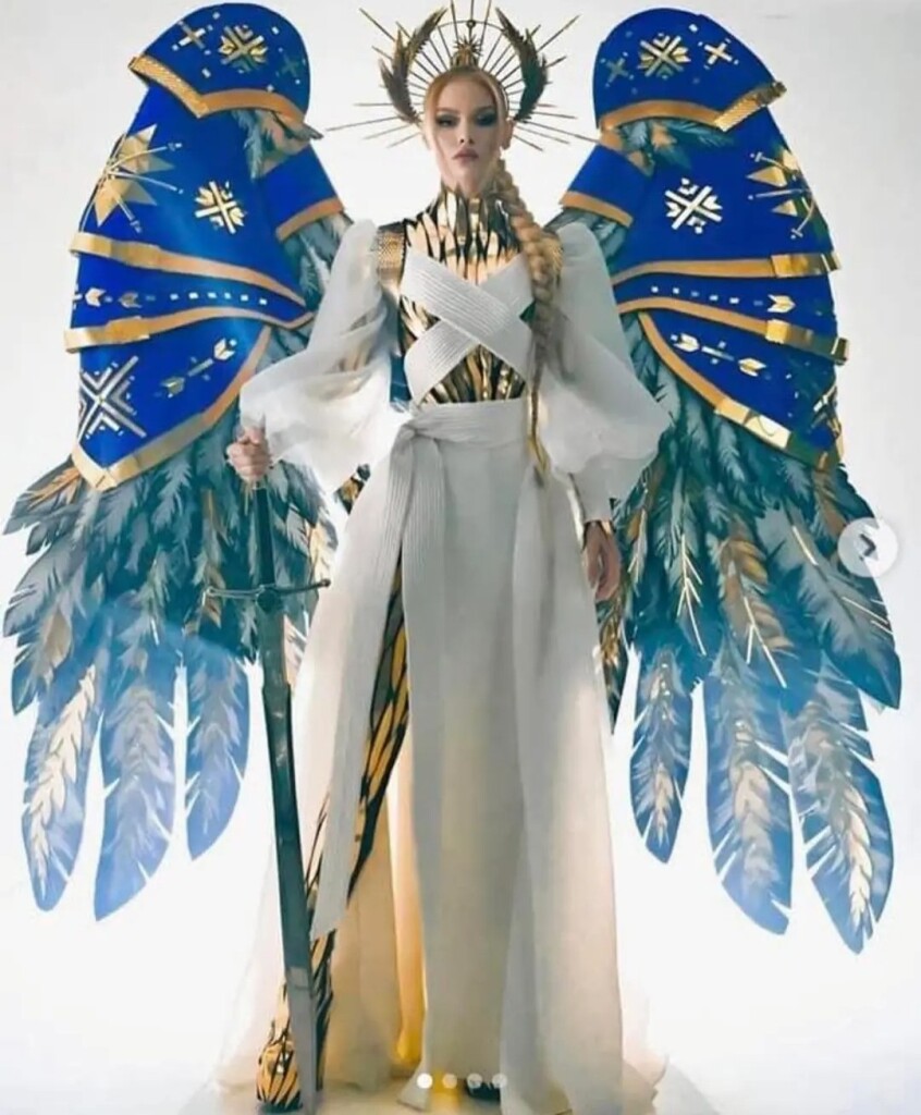 Miss Ukraine dressed as an angel with almost body length blue, white, and yellow feathers. The feathers are draped in the royal blue sash with golden symbols. Miss Ukraine is wearing a white dress with what looks like a body armor jumpsuit underneath. She is wearing a golden radiating crown, similar to what the Virgin Mary wears in artwork. She is also holding a 4 foot long sword, with the point digging into the ground. Her long blond braid cascades down her left shoulder. She looks stunningly fierce and defiant and truly captures the spirit of the Ukrainian people during this time of war with Russia. 