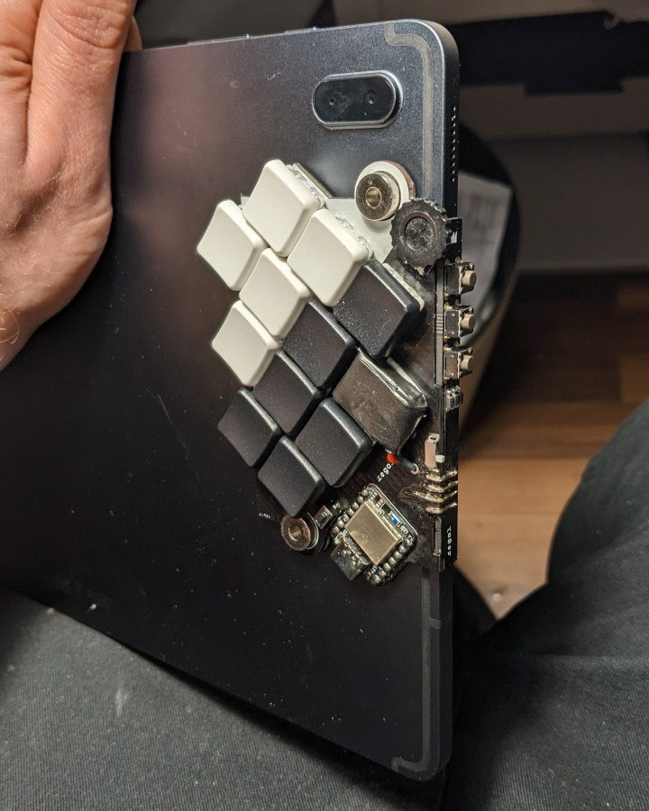 Photograph of a tablet computer's backside, resting on a person's lap. A small keyboard is stuck to the tablet's back on the right side. It adds some keys on the tablet's backside and some on the edge, with a 90° angle in-between. The keyboard itself is very flat. The 11 flat keys for the fingers are positioned such that each finger's length is taken into account. 3 very small buttons on the side are reachable with the thumb. A small flat scroll wheel is positioned in the top corner.