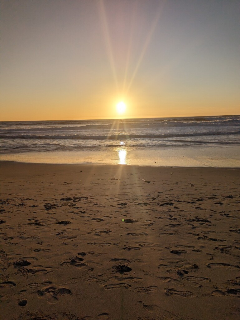 The sun setting over the ocean, with a sandy shore in the foreground. Rays of light spread out on the lens.