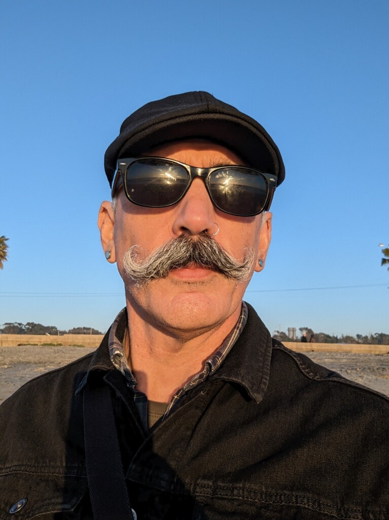 Evan Prodromou, in a black cap, knockoff Raybans, a big gray handlebar moustache, a brown plaid shirt and a black jeans jacket, faces into the golden light of the sunset. Behind, palm trees and sand, with cypress in the far distance.