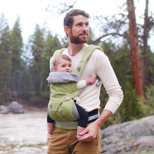 A white European man stands in a forest clearing, with a baby in a baby Bjorn style carrier. The man is smiling. Because he's holding a baby. Babies are cute.