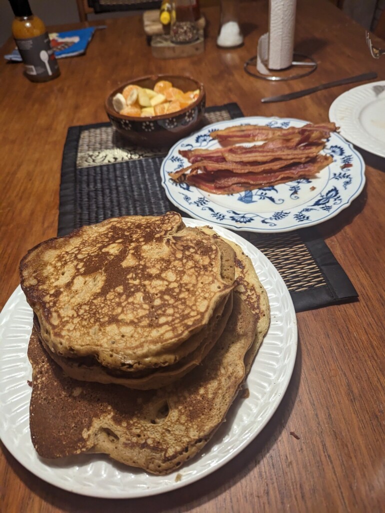 A stack of pancakes on a white plate on a wooden table. Behind them, a plate of bacon. Behind that, a fruit salad.
