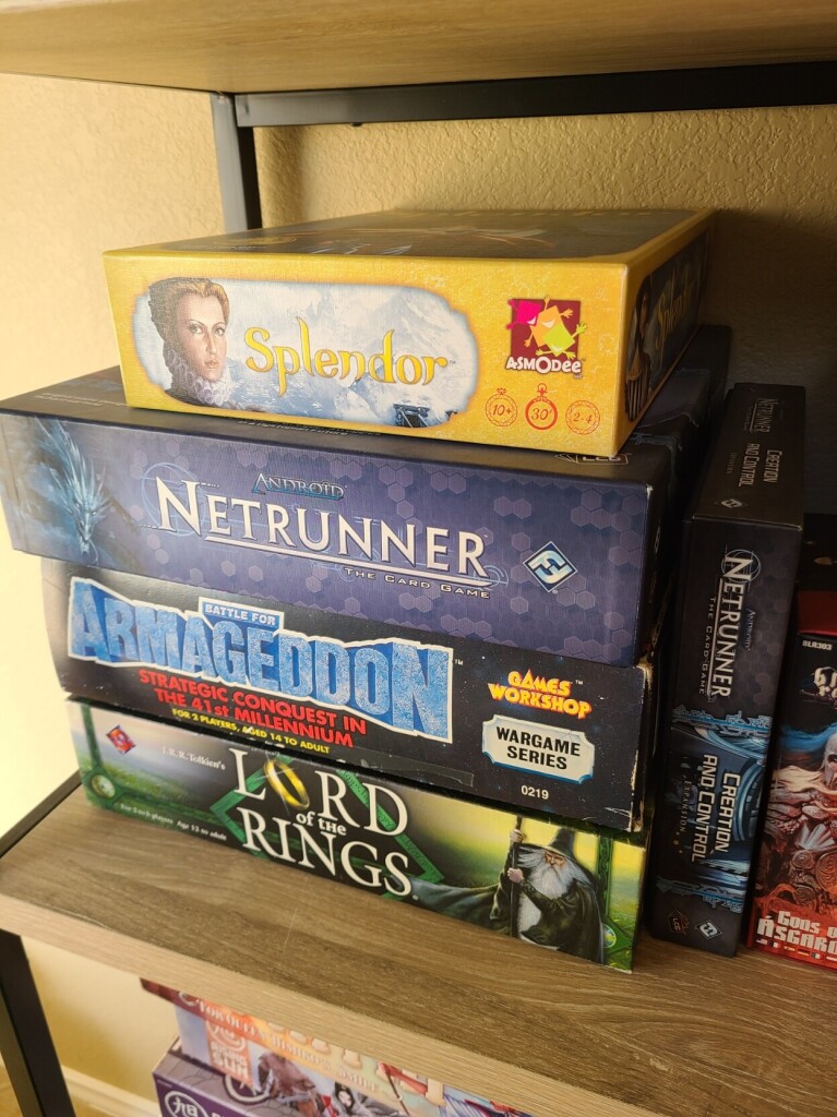 A stack of games sitting on a shelf. The games are Splendor, Netrunner, Netrunnet: Command & Control expansion, Battle for Armageddon, and Lord of the Rings.