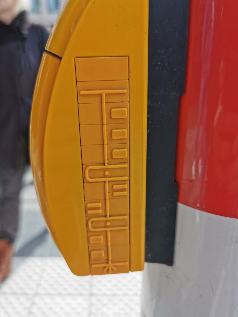 A button at a pedestrian crosswalk, viewed from the side. There is an embossed diagram of the crossing's layout with a line indicating the path you walk along and several rectangular pieces indicating car lanes and tram tracks. 