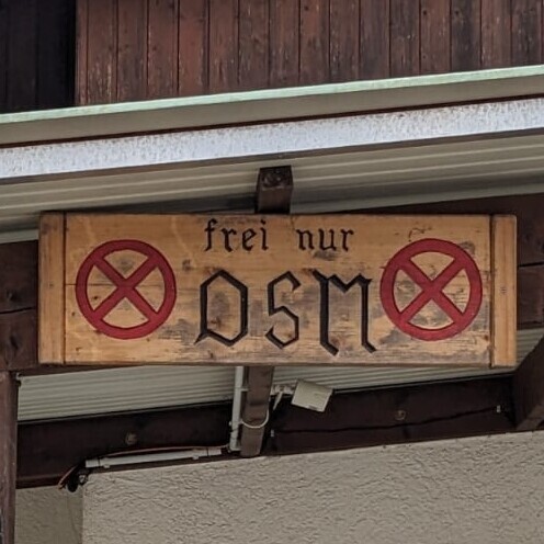 Wooden sign, with a red crossed circle on the left and right. Text , in old German script says "frei nur / OSM"